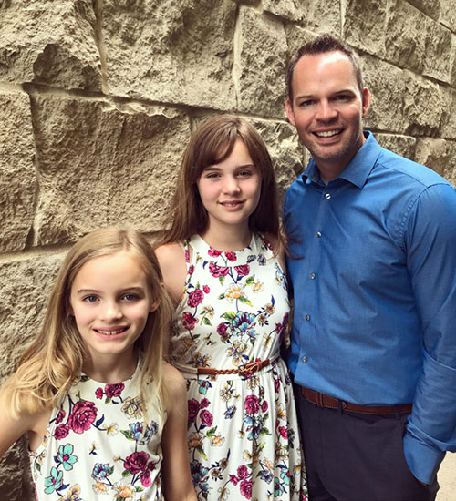 Tony standing with his two daughters who are wearing matching flowery dresses